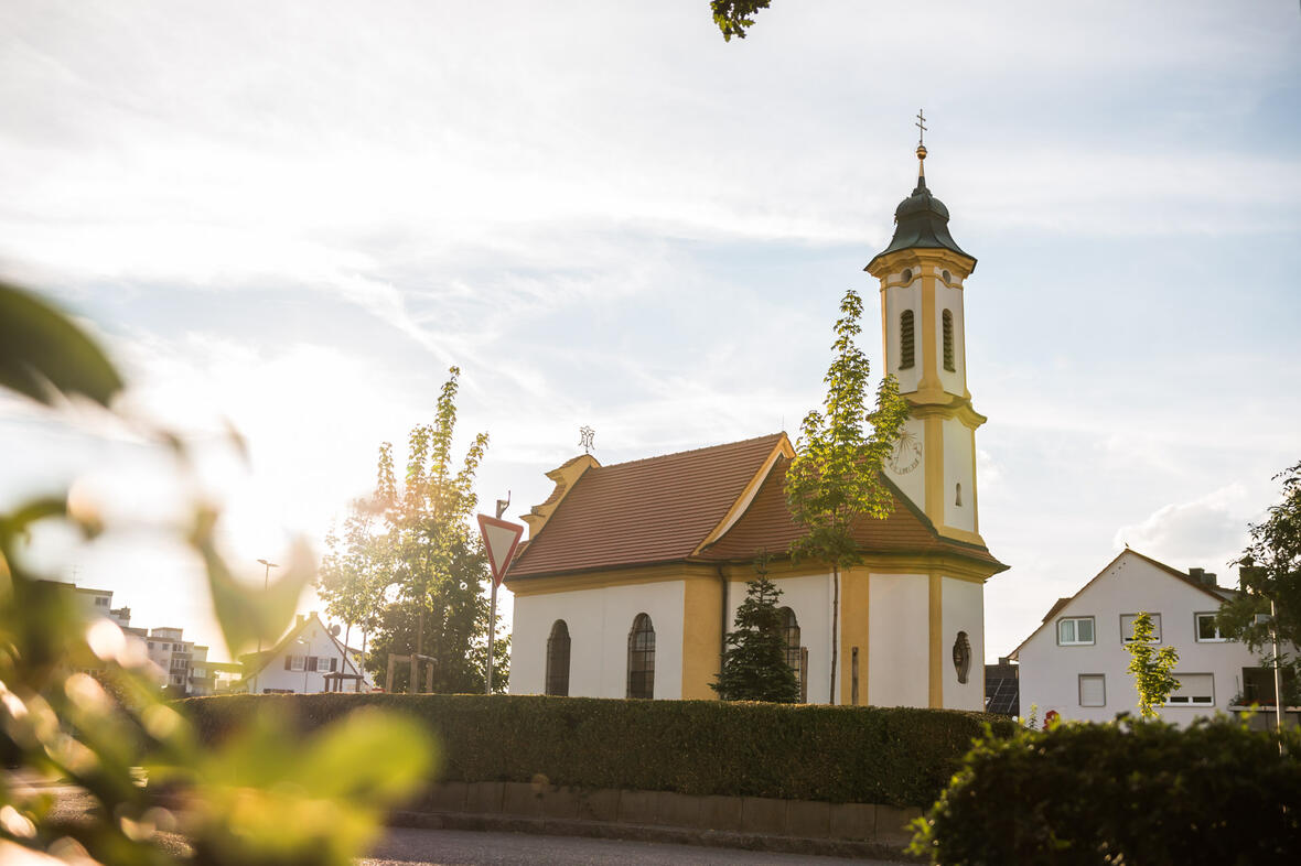 Die Kapelle im Morgenlicht, umgeben von Bäumen und einem gepflegten Grünstreifen.