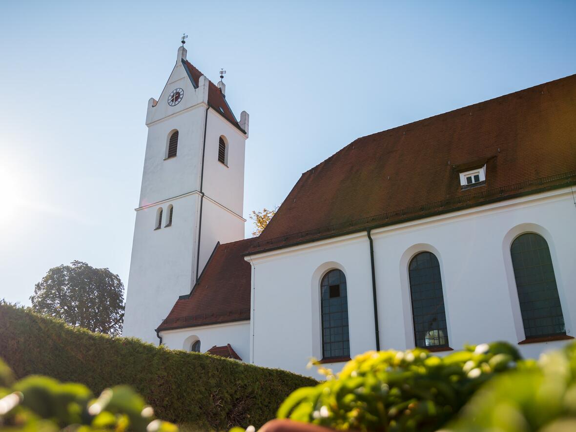 Kirche mit markantem Turm und weißen Wänden, umgeben von grüner Vegetation im Sonnenlicht.