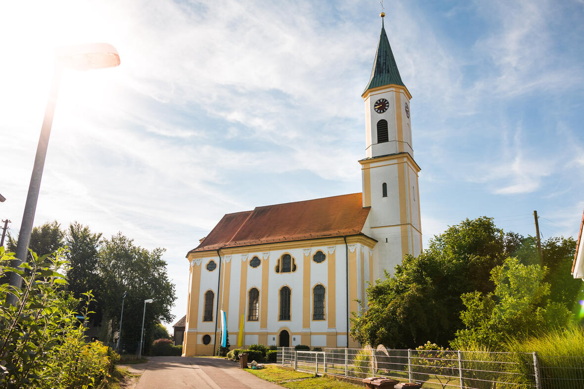 Kirche in ruhiger Umgebung, im Sonnenlicht, umgeben von Bäumen und einem Feldweg.