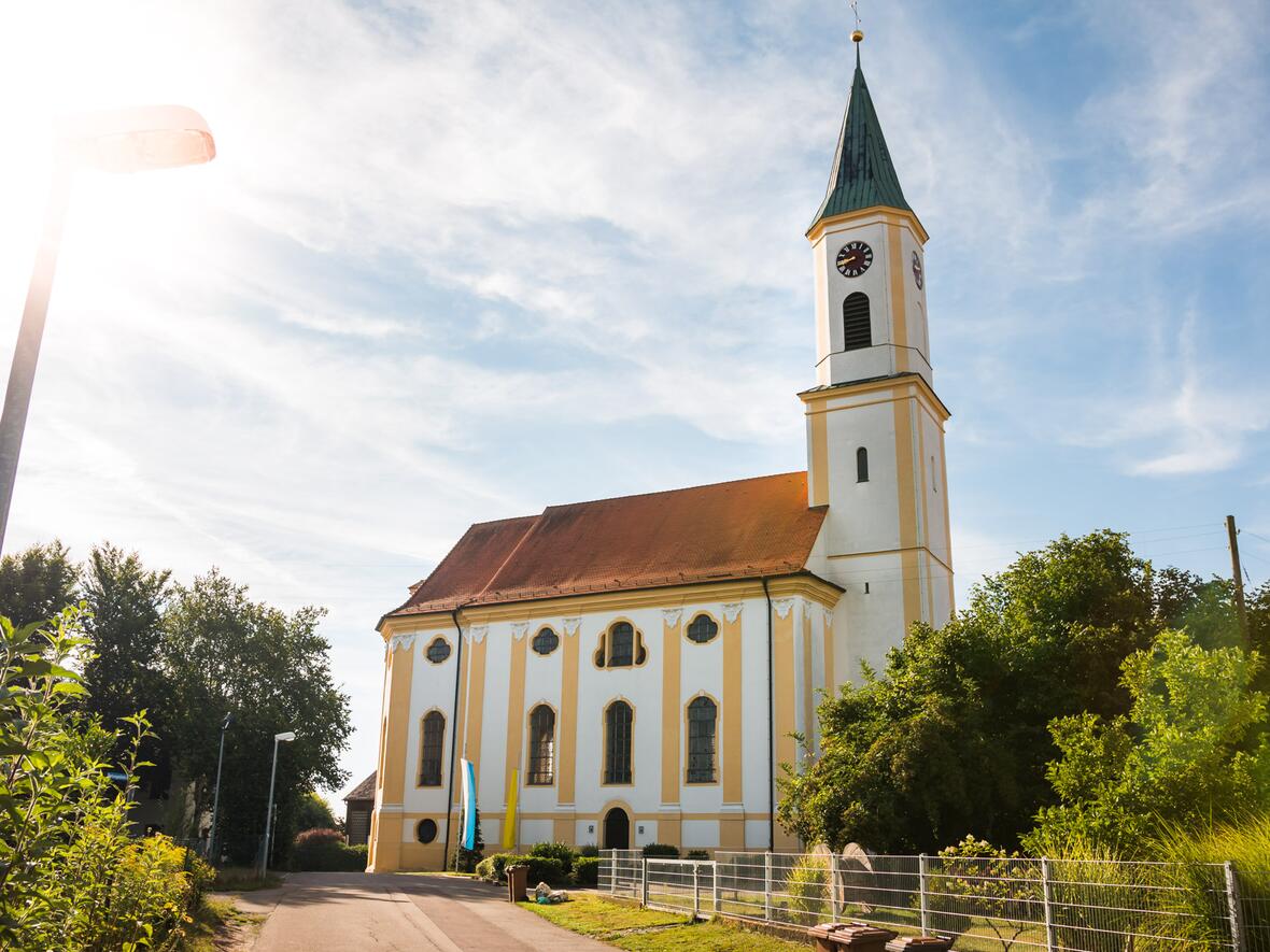 Kirche in ruhiger Umgebung, im Sonnenlicht, umgeben von Bäumen und einem Feldweg.