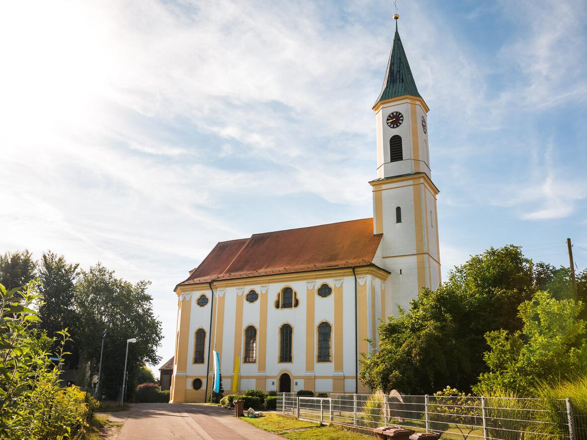 Kirche in ruhiger Umgebung, im Sonnenlicht, umgeben von Bäumen und einem Feldweg.